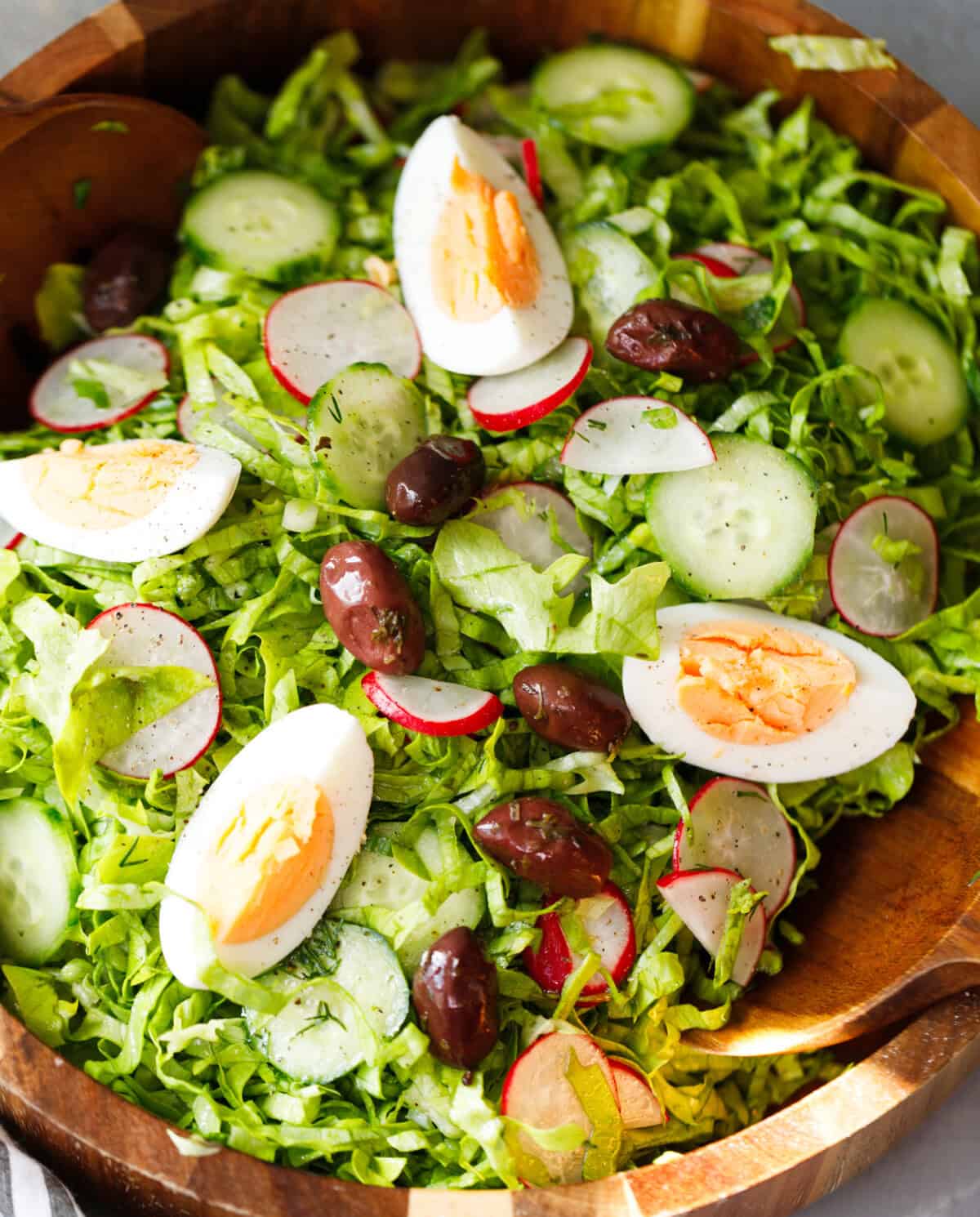Bulgarian green salad with lettuce, cucumbers, radishes, eggs and olives in a wooden bowl with serving spoon