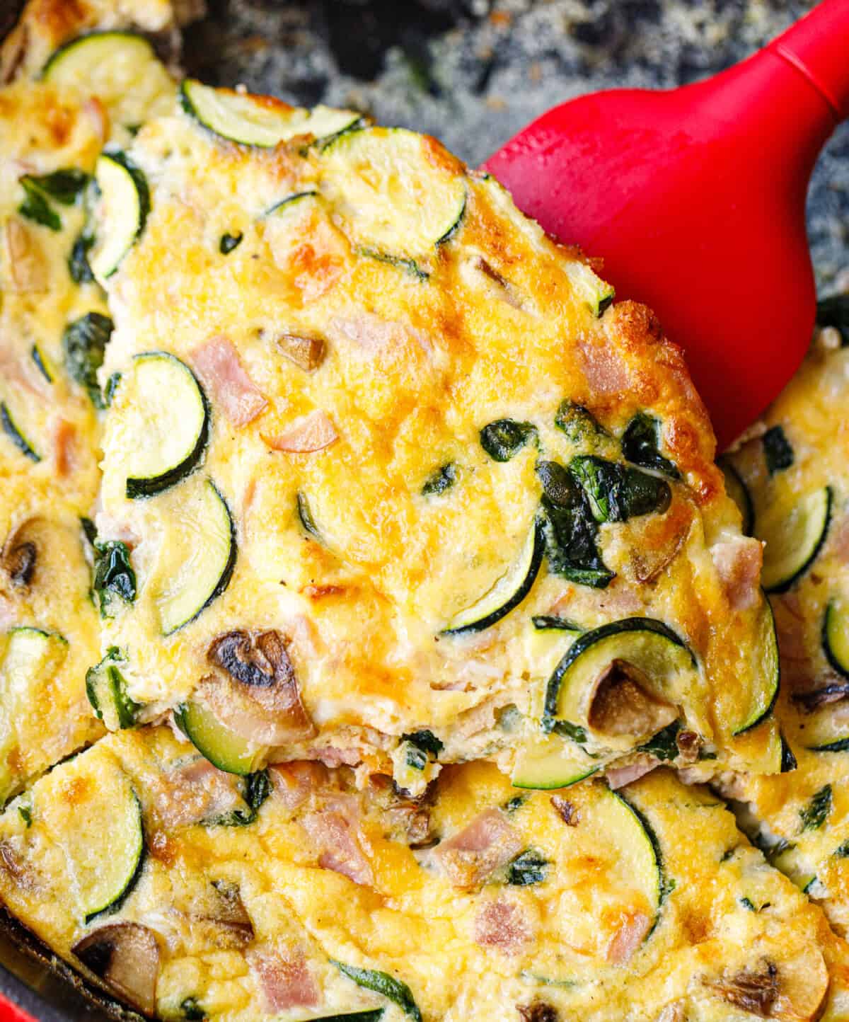 Red spatula lifting a slice of spinach mushroom frittata from a cast iron skillet