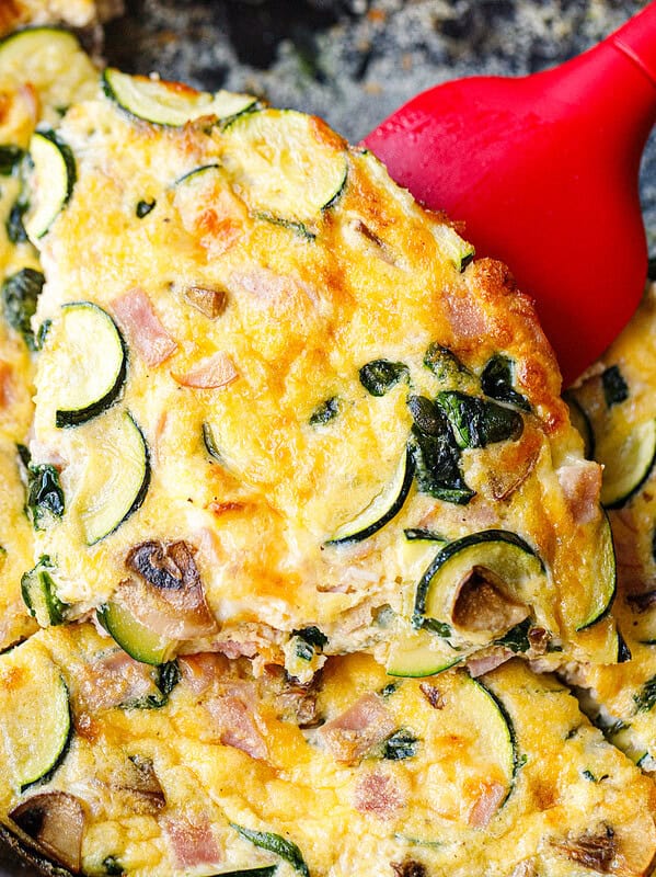 Red spatula lifting a slice of spinach mushroom frittata from a cast iron skillet