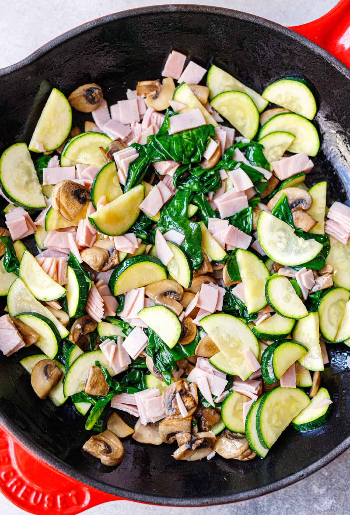 Cast iron skillet with sautéed zucchini, mushrooms, wilted spinach, and chopped ham for a spinach mushroom frittata