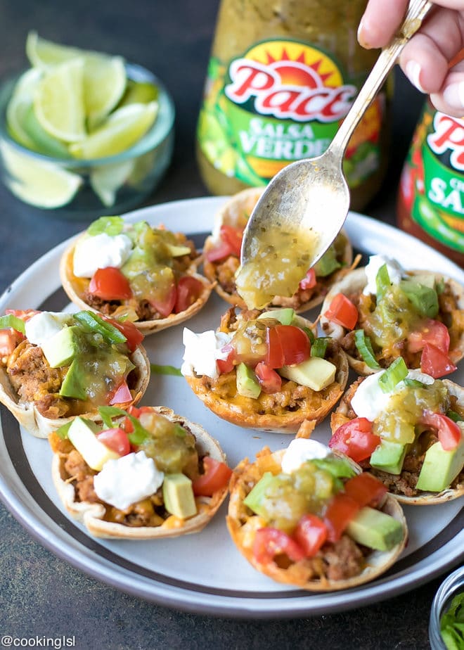 Crunchy Taco Cups With Tortillas, turkey and salsa verde recipe. Made with Pace® Salsa and perfect for a game day appetizer. A plate with mini taco appetizer, topped with avocado, sour cream and tomatoes. Lime wedges on the side. Topped with Pace Salsa Verde.