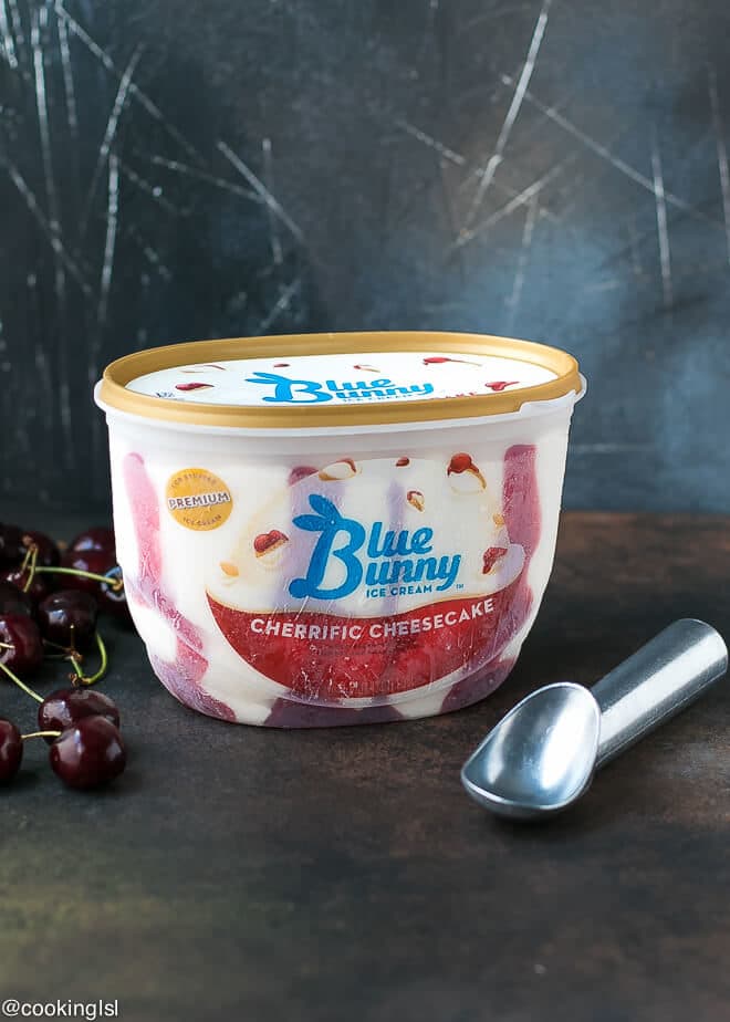Cherry Sundae Recipe, made with Blue Bunny® Ice Cream, Fresh fruit, cookie crumbs and nuts. A container of Blue Bunny Cherrific Cheesecake Ice cream with a metal ice cream scoop next to it.