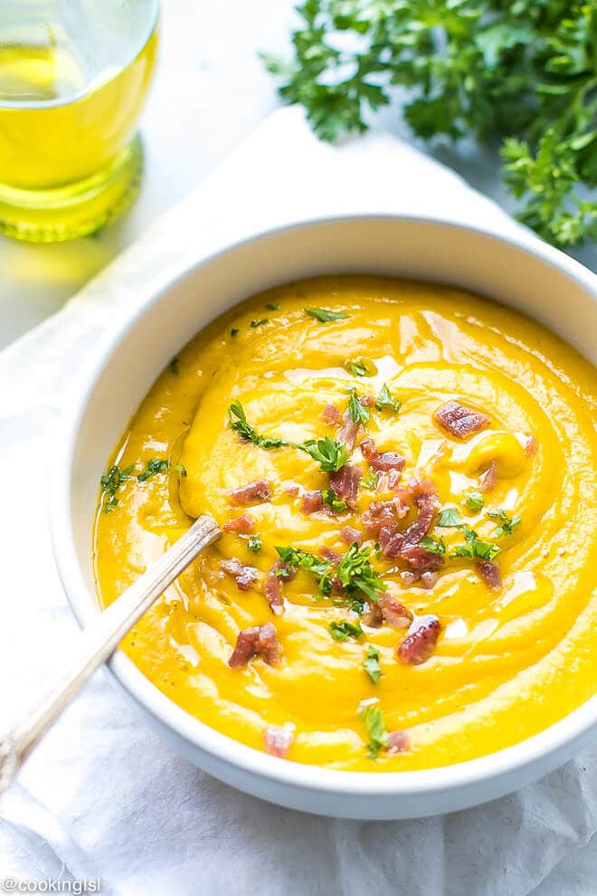 A white creamic bowl with creamy sweet potato soup, rich orange color and topped with bacon bits and chopped parsley.