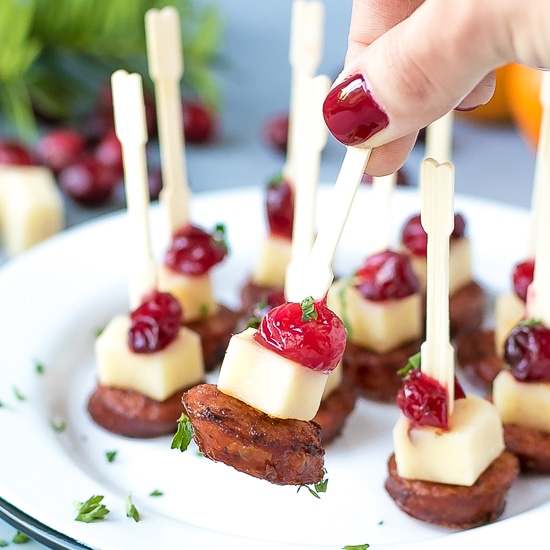 Andouille Sausage Appetizer Bites With Cranberry Cheddar - mini bites on a plate with wooden skewers, held in hand.