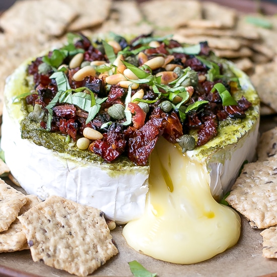 Savory Baked Brie Appetizer Recipe. Brie wound on a platter, topped with pesto, sun dried tomatoes, pine nuts, basil and crackers next to it.