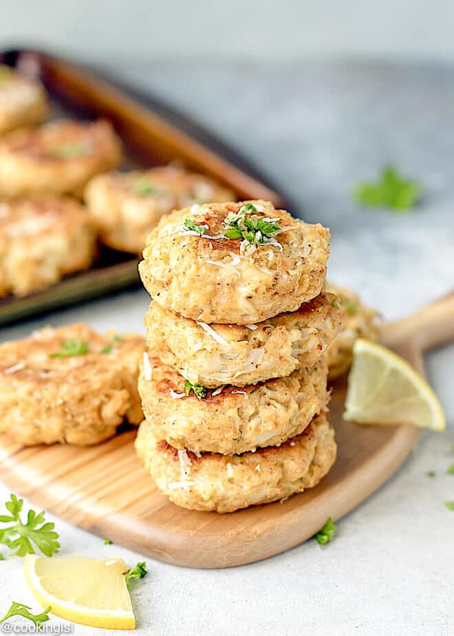 Cheesy cauliflower patties on top of each other on a wooden board with lemon slice on the side