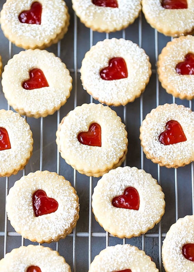 Easy Almond Linzer Cookies Recipe, round cutout cookies, sandwiched together with strawberry jam, placed on a drying rack.
