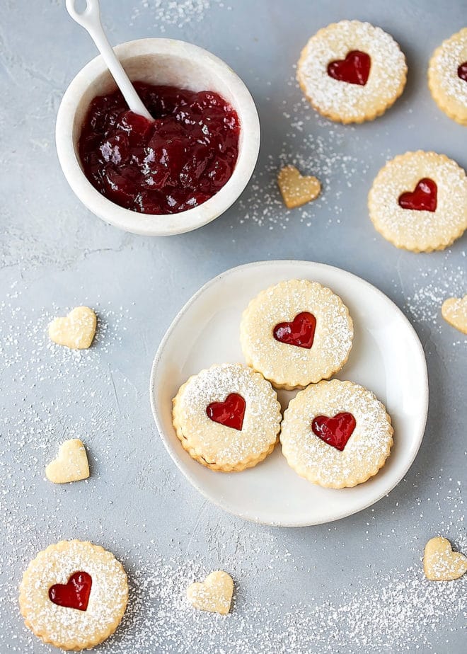 Easy Almond Linzer Cookies Recipe, round cutout cookies, sandwiched together with strawberry jam, dusted with powdered sugar, placed on a small round white plate. Small white bowl filled with jam on the side. Heart cutouts, great for Valentine's Day.