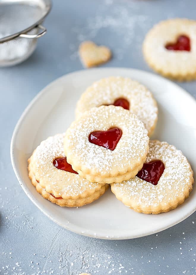 Easy Almond Linzer Cookies Recipe, round cutout cookies, sandwiched together with strawberry jam, dusted with powdered sugar, placed on a small round white plate.