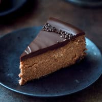 Chocolate Mascarpone Cheesecake Recipe. A slice of chocolate cheesecake topped with thick chocolate ganache. The perfect slice on a black plate.