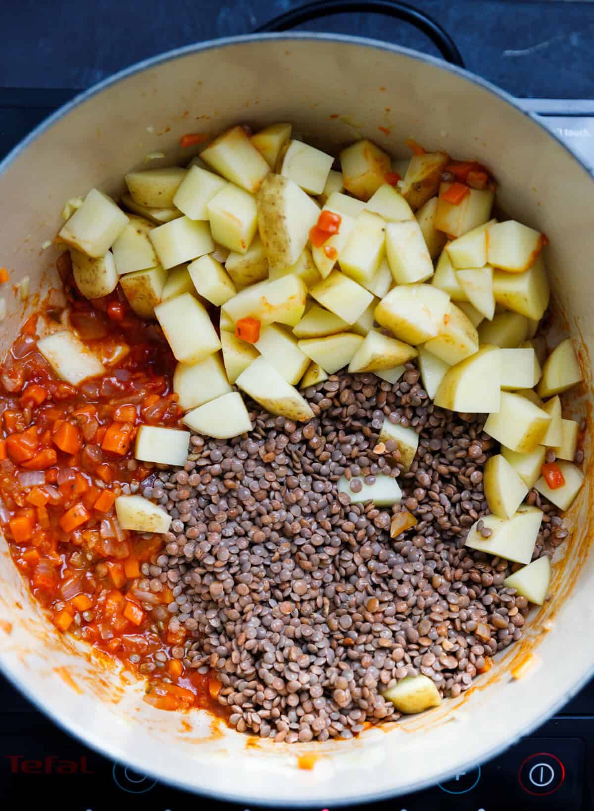 A Dutch oven with onions, carrots, garlic, tomato sauce and paste and cubbed potatoes inside.