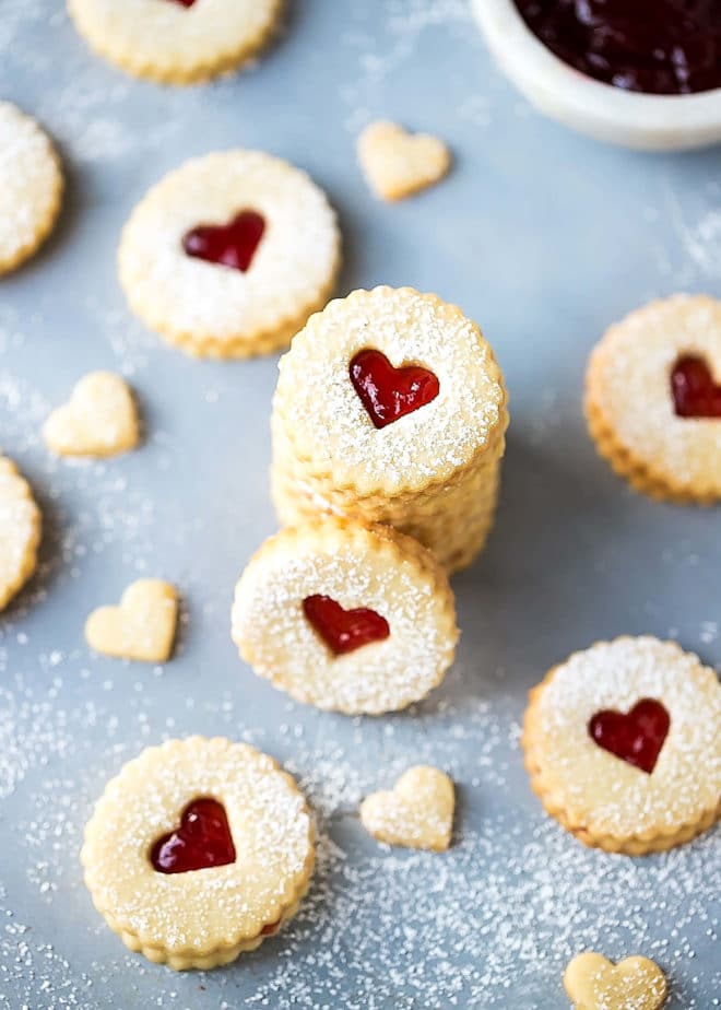 Easy Almond Linzer Cookies Recipe, round cutout cookies, sandwiched together with strawberry jam, dusted with powdered sugar, placed on a small round white plate. Small white bowl filled with jam on the side. Heart cutouts, great for Valentine's Day. Stacked on top of each other.
