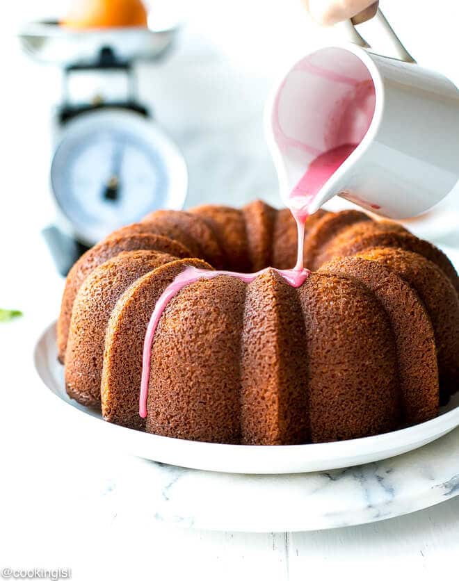 Blood Orange Bundt Cake- LOADED WITH CITRUS FLAVOR, ON A WHITE PLATE, WITH PINK GLAZE POURING OVER IT.