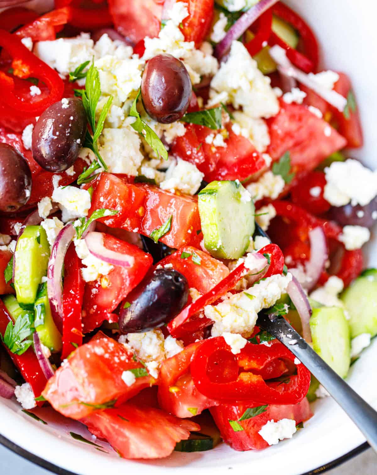  Close up of shopska salad with tomatoes, cucumbers, red peppers, kalamata olives, and grated feta in a white enamel bowl