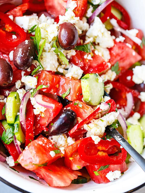 Close up of shopska salad with tomatoes, cucumbers, red peppers, kalamata olives, and grated feta in a white enamel bowl