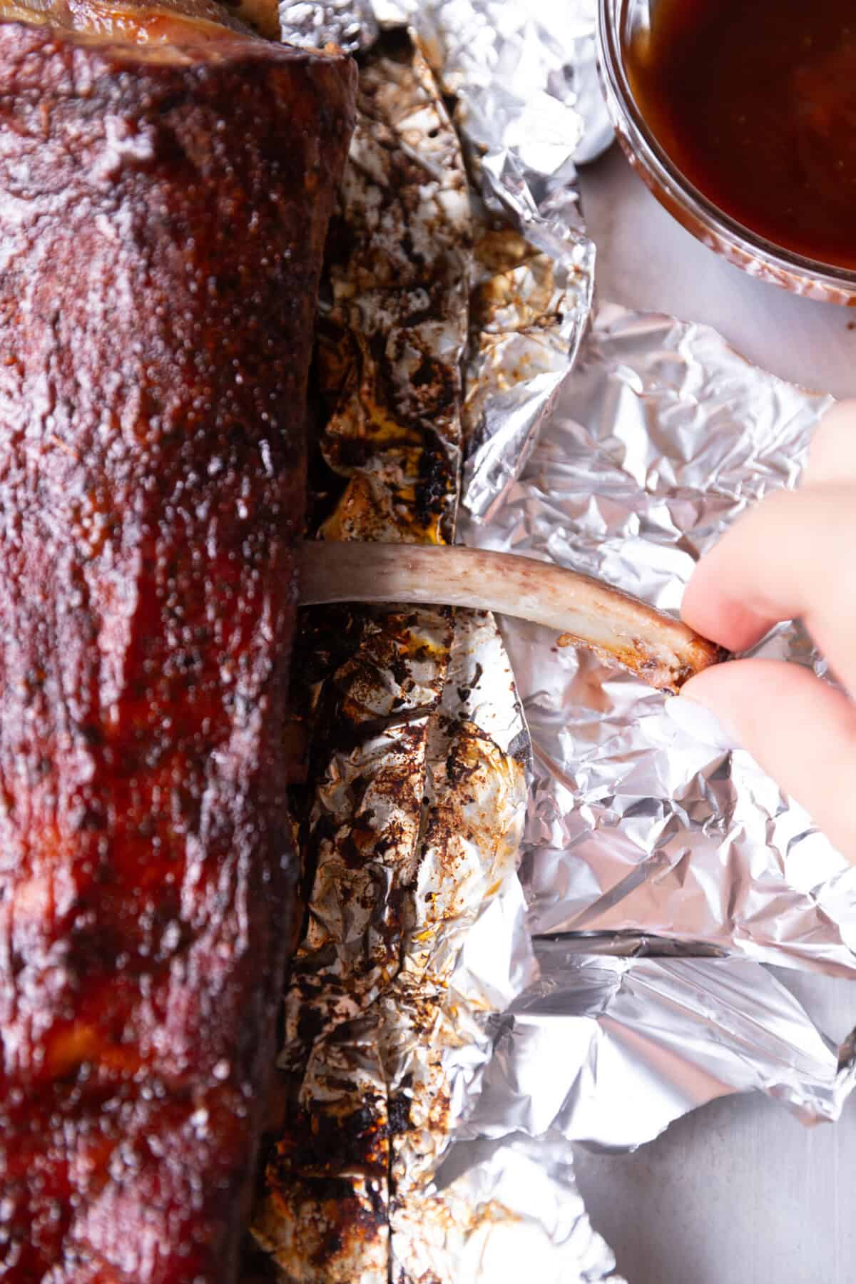 Pulling a tender oven-baked St. Louis style rib from a foil-lined baking sheet after cooking, with barbecue sauce and charred edges visible.