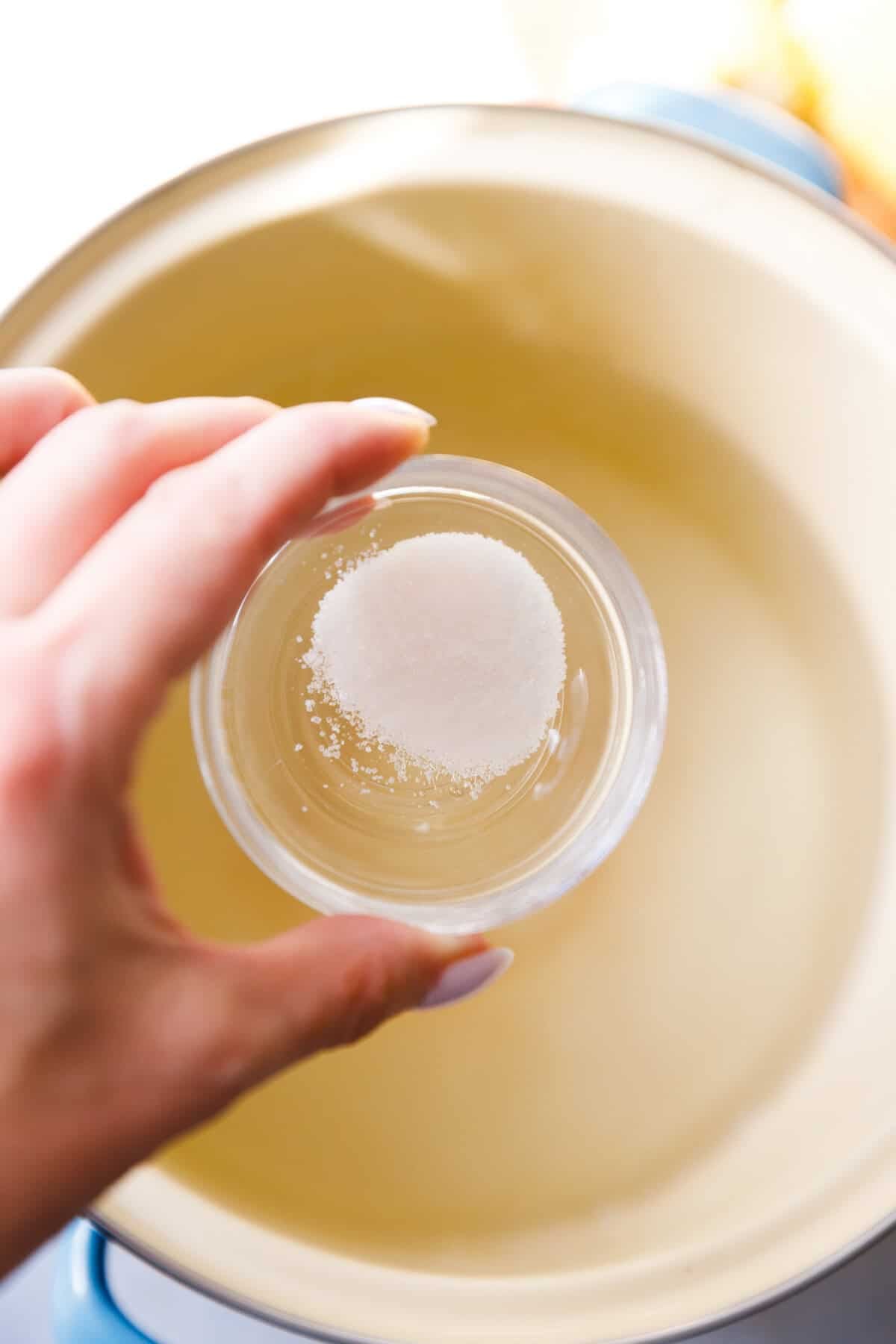 A hand holding small bowl with sugar over a pot