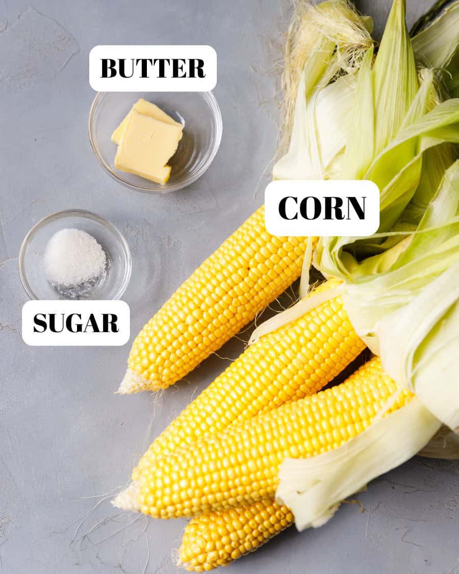 Ingredients for boiled corn on the cob: fresh yellow corn, butter, and sugar on a gray background.