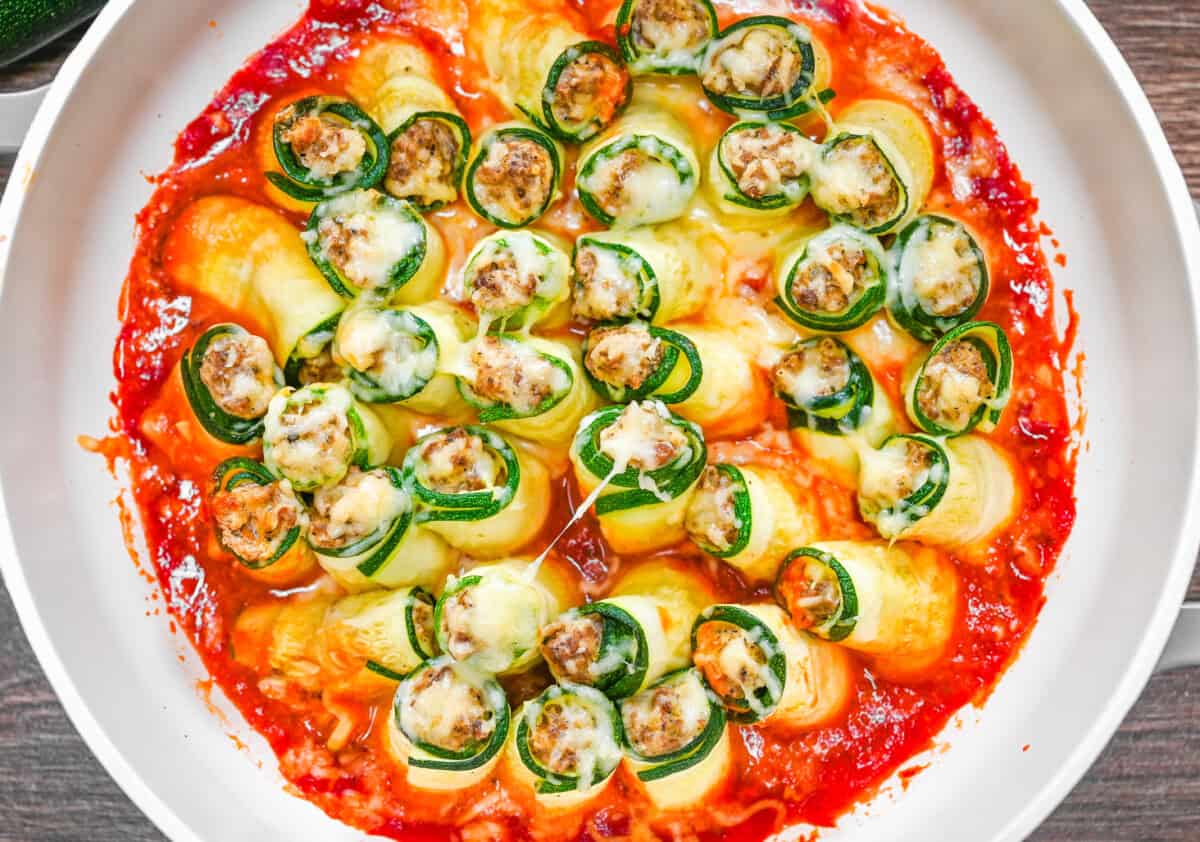 Overhead view of baked zucchini lasagna roll ups in a white skillet, topped with melted mozzarella and tomato sauce.