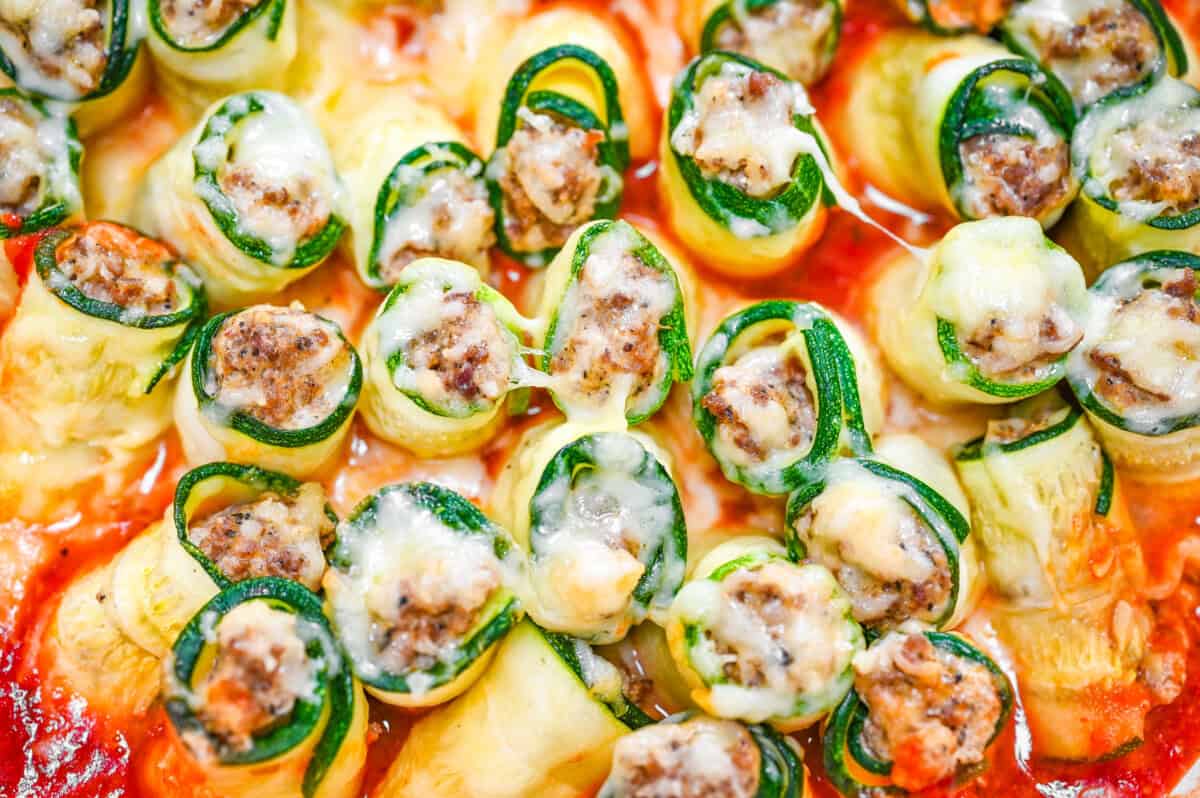 Close-up of baked zucchini lasagna roll-ups filled with ground meat and topped with melted cheese over a layer of tomato sauce.