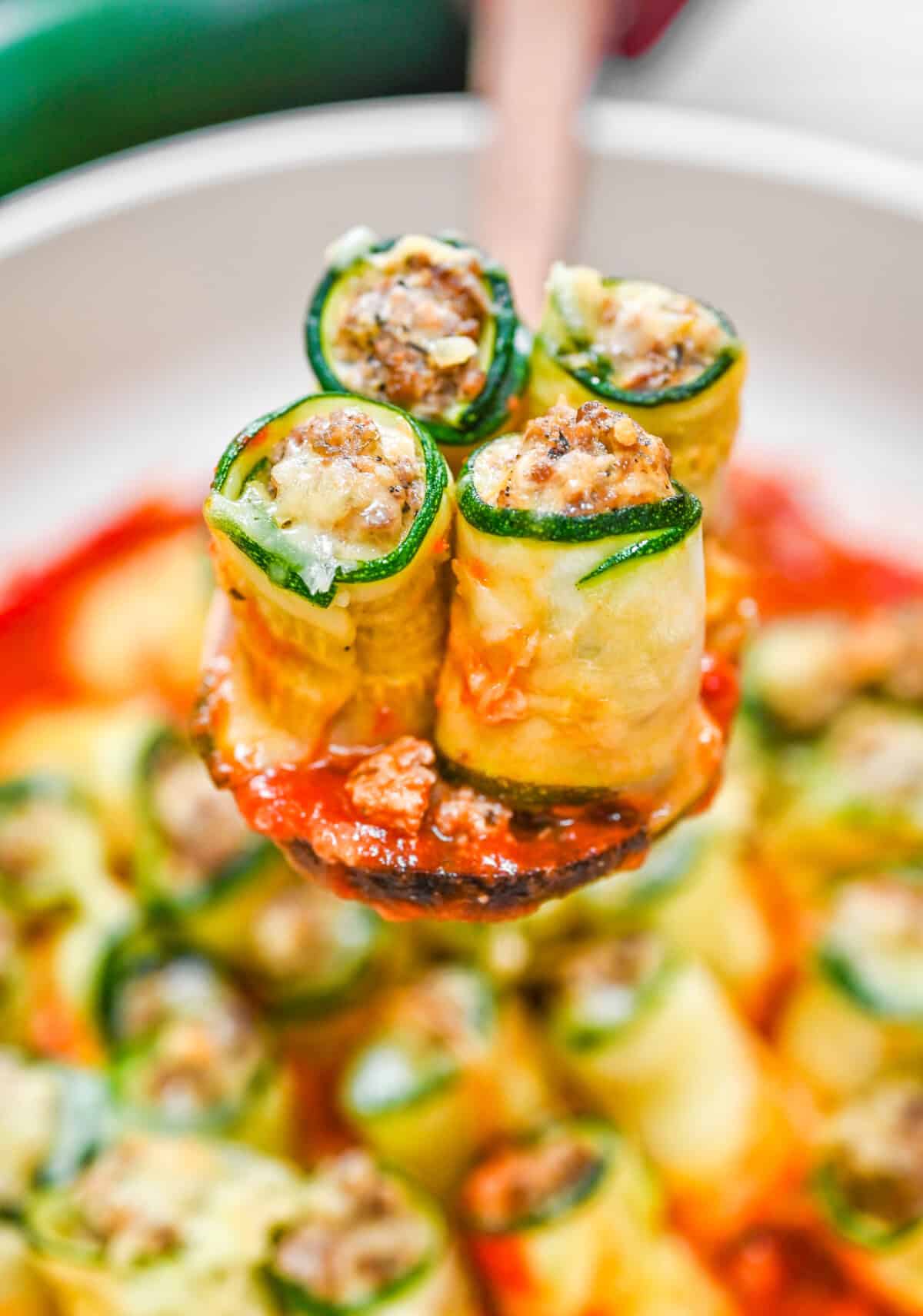 Close-up of zucchini lasagna roll ups filled with ground beef and cheese, lifted from a skillet with marinara sauce underneath.