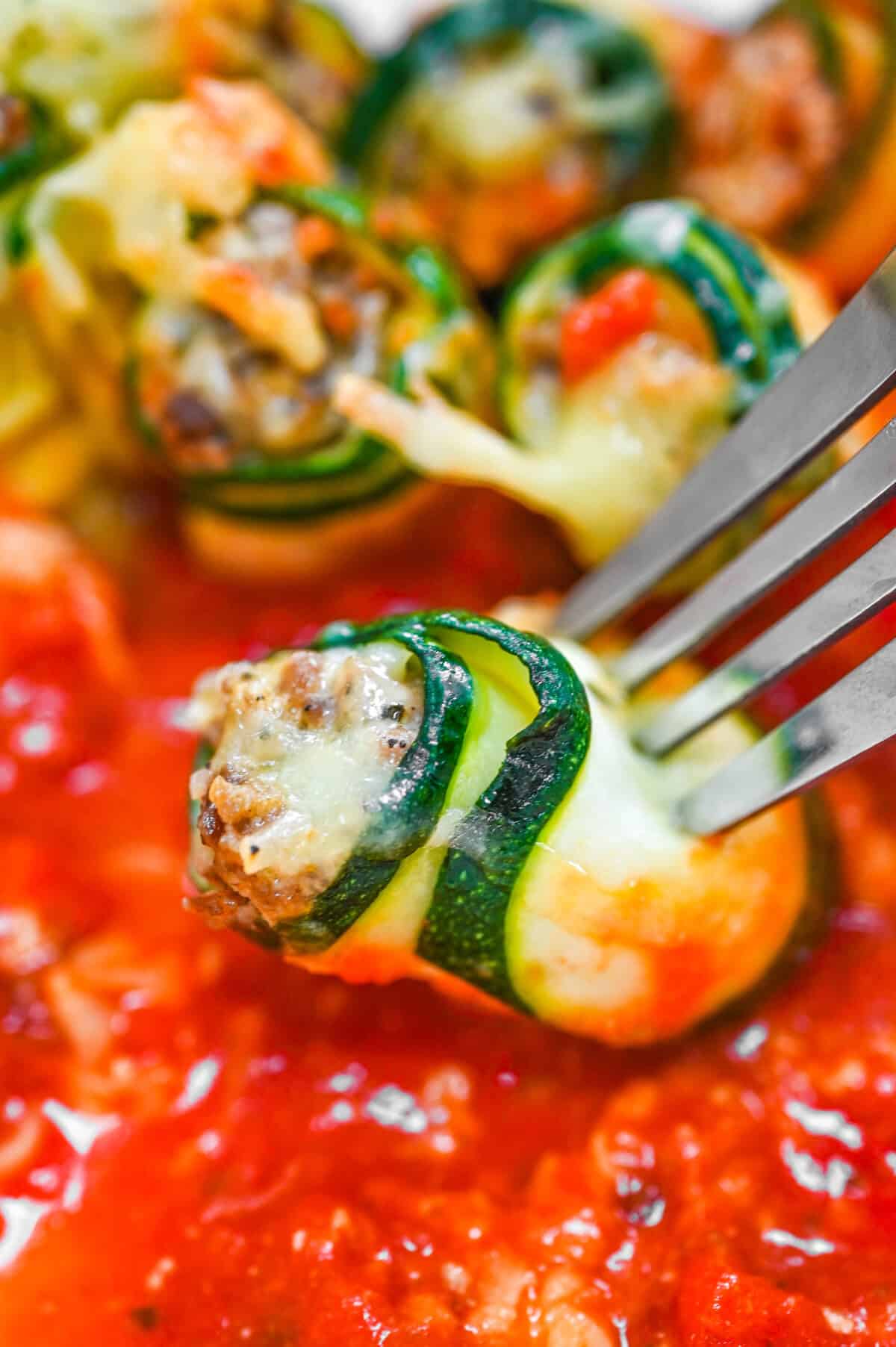 Close-up of a cheesy zucchini lasagna roll-up with meat filling on a fork, lifted above a rich tomato sauce.


