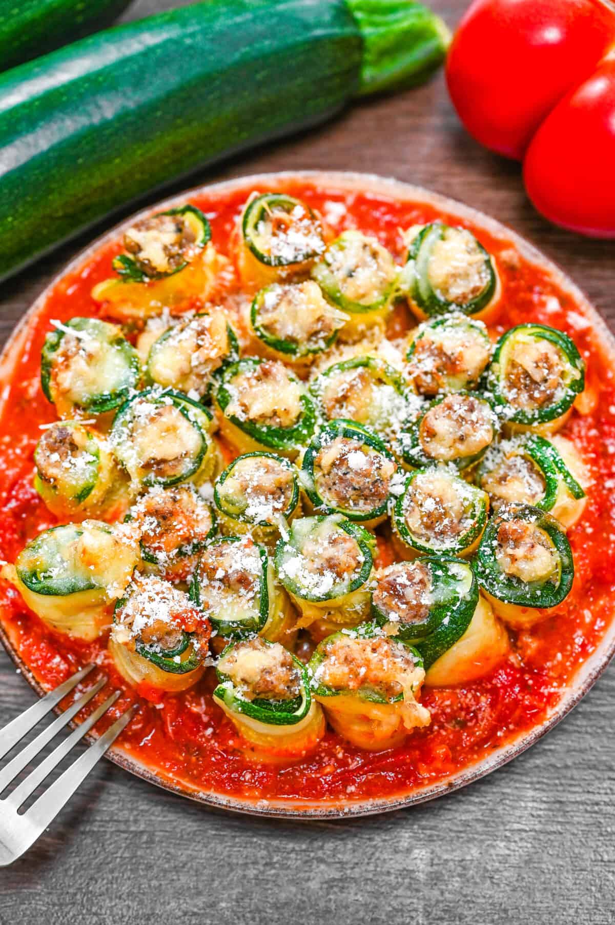 A plate of zucchini lasagna roll-ups topped with cheese and Parmesan, surrounded by tomato sauce, with fresh zucchini and tomatoes in the background.
