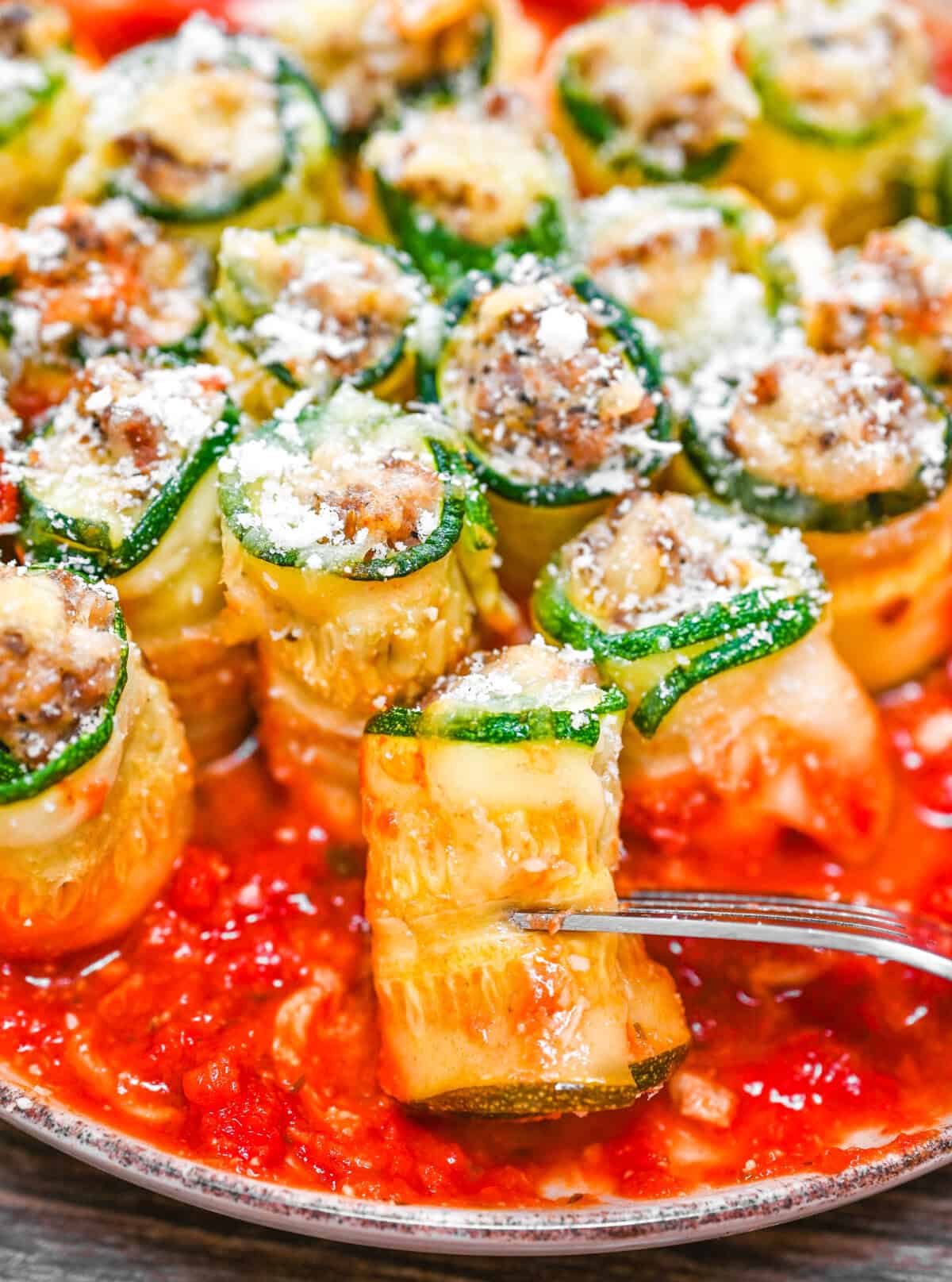 Close-up of a cheesy zucchini roll-up on a fork, surrounded by sauce and topped with Parmesan and beef filling.