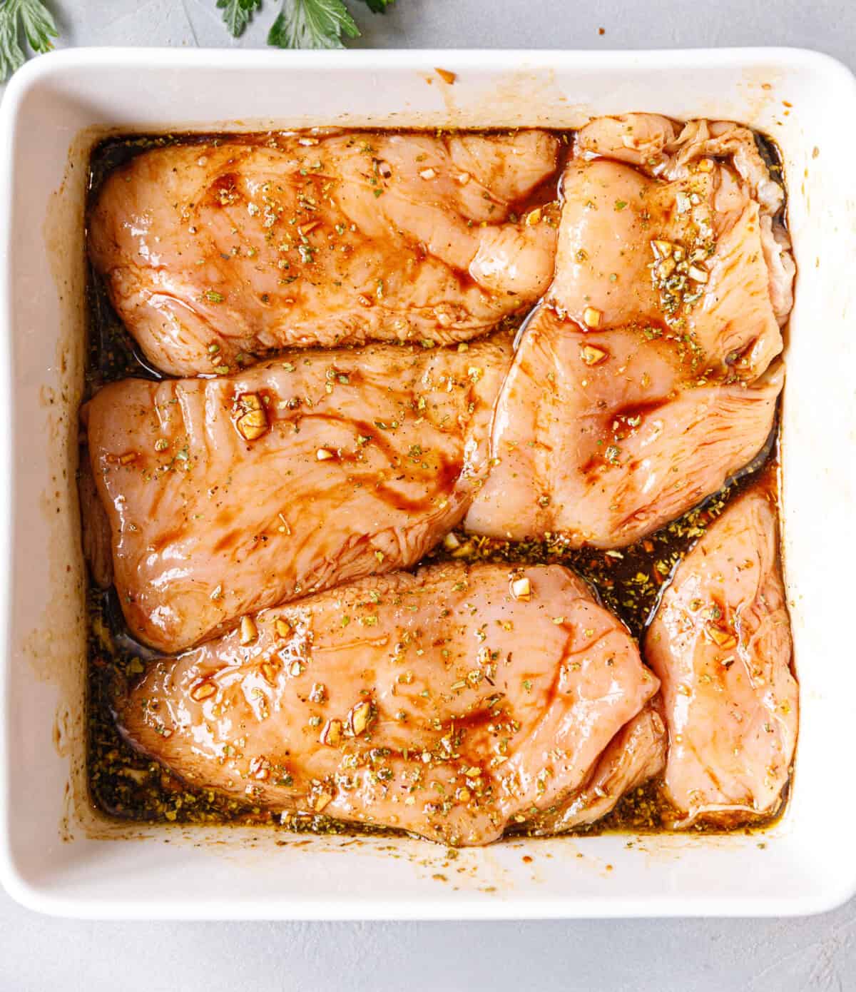 Raw chicken breasts coated in balsamic, garlic, and herb marinade in a white baking dish, ready to bake.