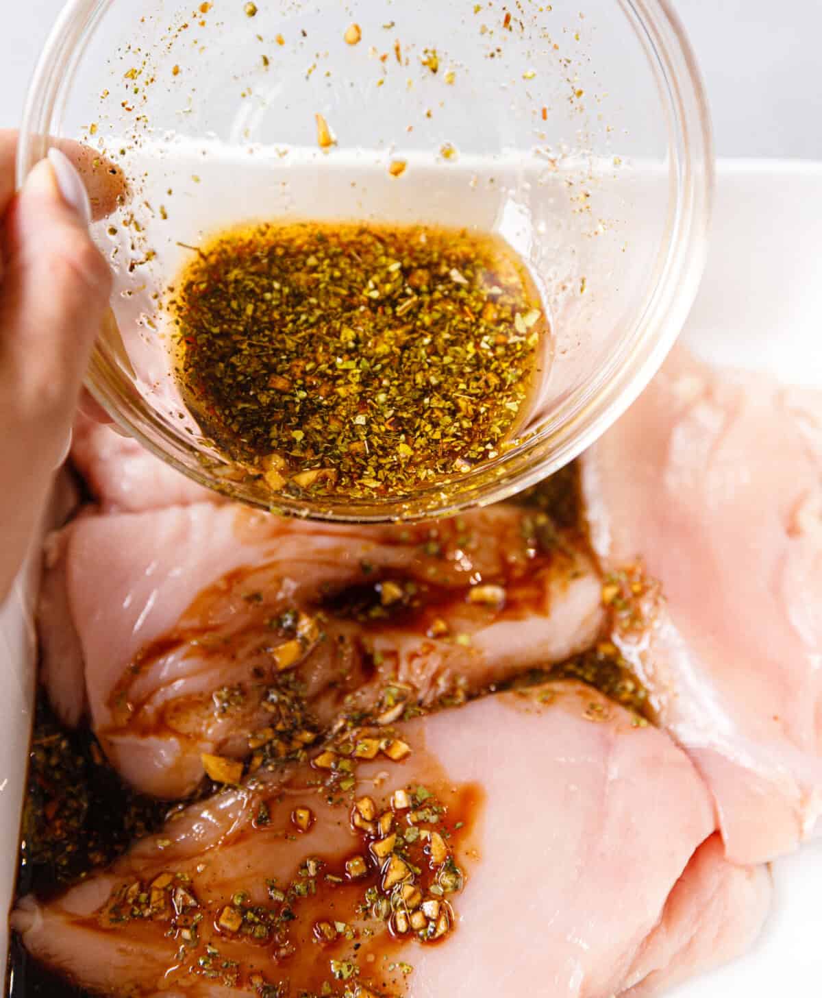 Pouring homemade balsamic marinade with garlic and oregano over raw chicken breasts in a white baking dish before baking.