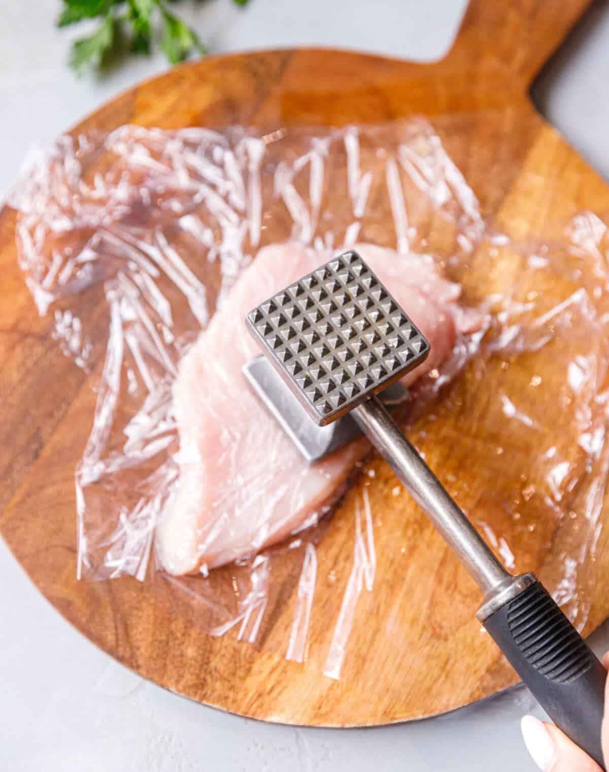 Flattening raw chicken breast under plastic wrap with a metal meat mallet on a wooden board to make it tender and even for baking.