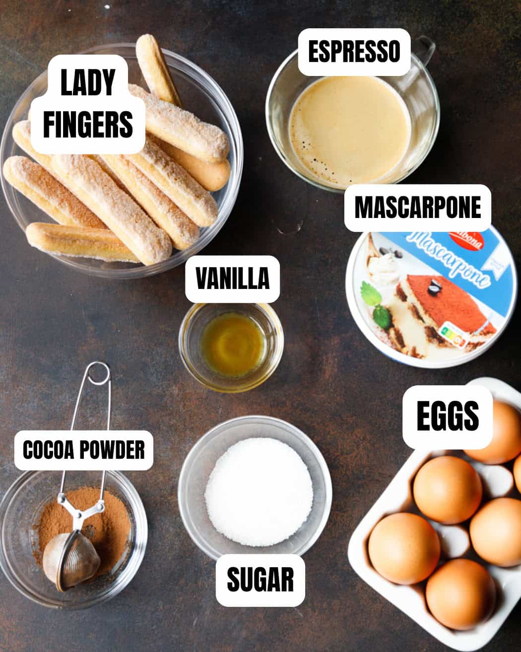 Overhead view of classic tiramisu ingredients in bowls, including ladyfingers, espresso, mascarpone, eggs, sugar, cocoa powder, and vanilla.