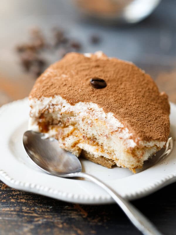 Close-up slice of classic tiramisu dusted with cocoa powder, served on a white plate with a spoon.