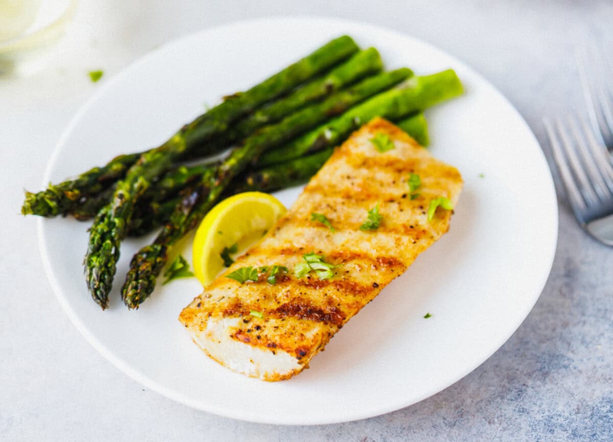 Grilled mahi mahi fillet with grill marks on a white plate with grilled asparagus and lemon wedge