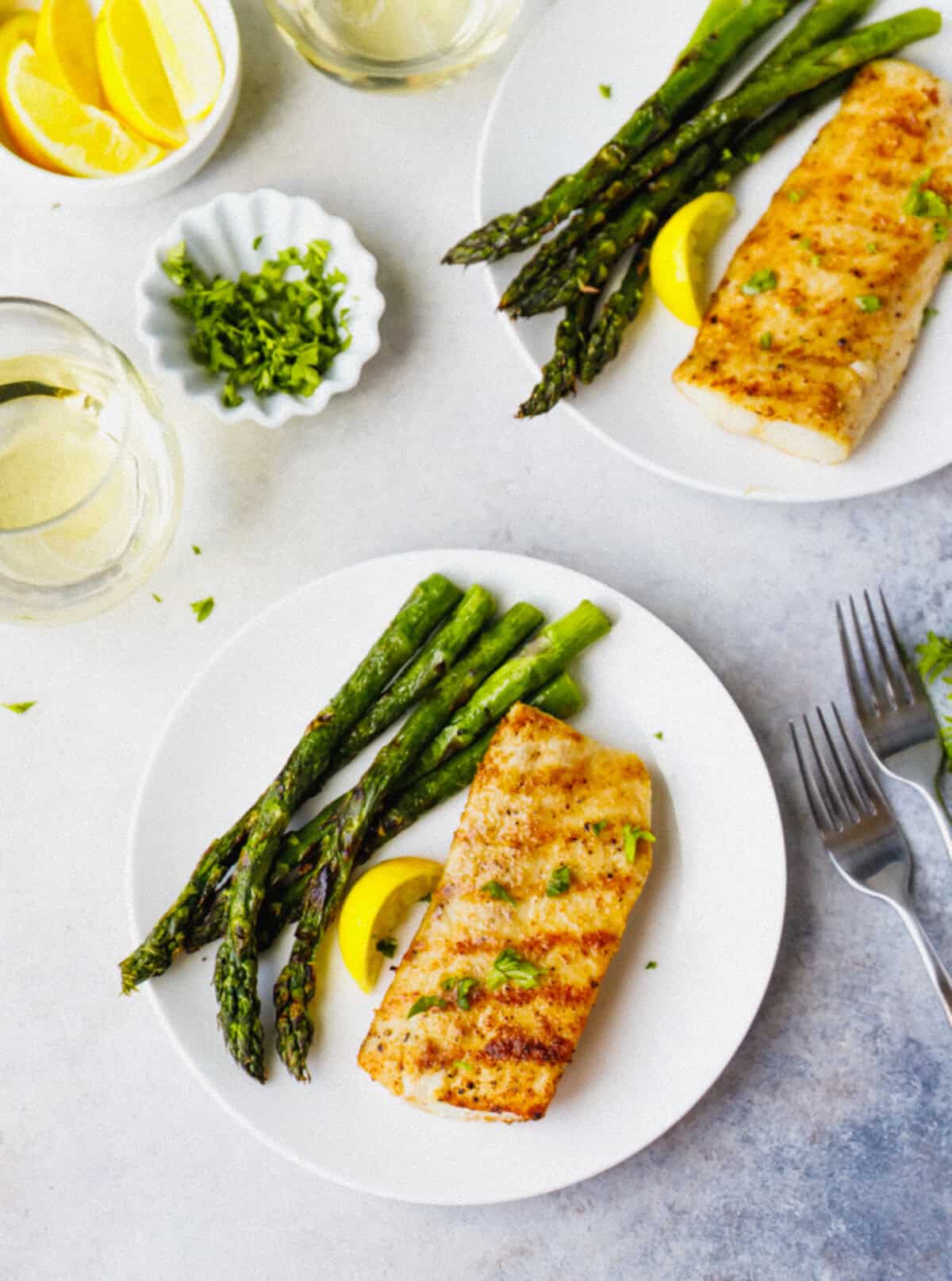 Overhead view of two plates with grilled mahi mahi and asparagus, white wine, and fresh parsley