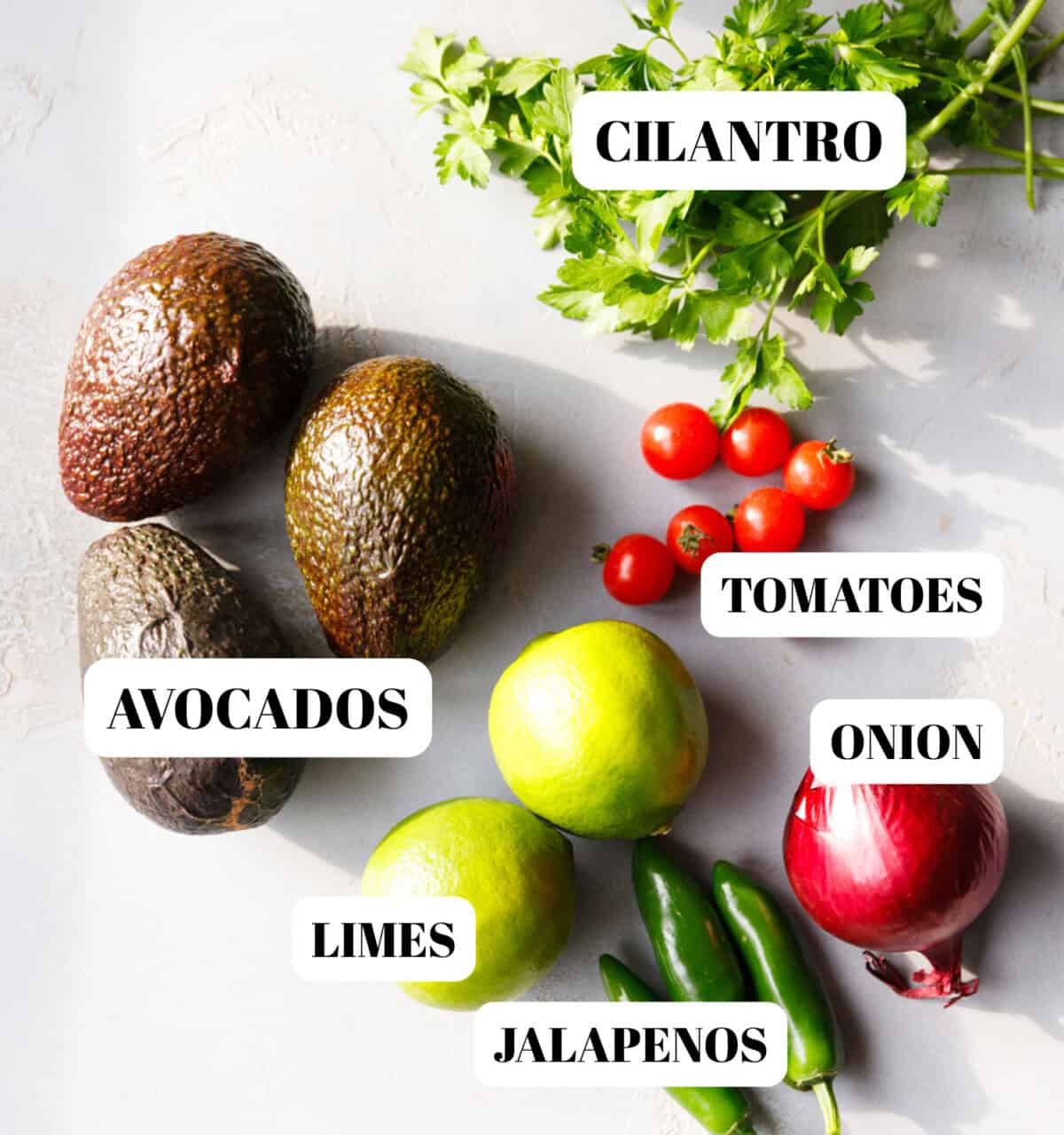 Fresh guacamole ingredients on a light surface including avocados, cilantro, cherry tomatoes, red onion, limes, and jalapeños labeled for easy identification.