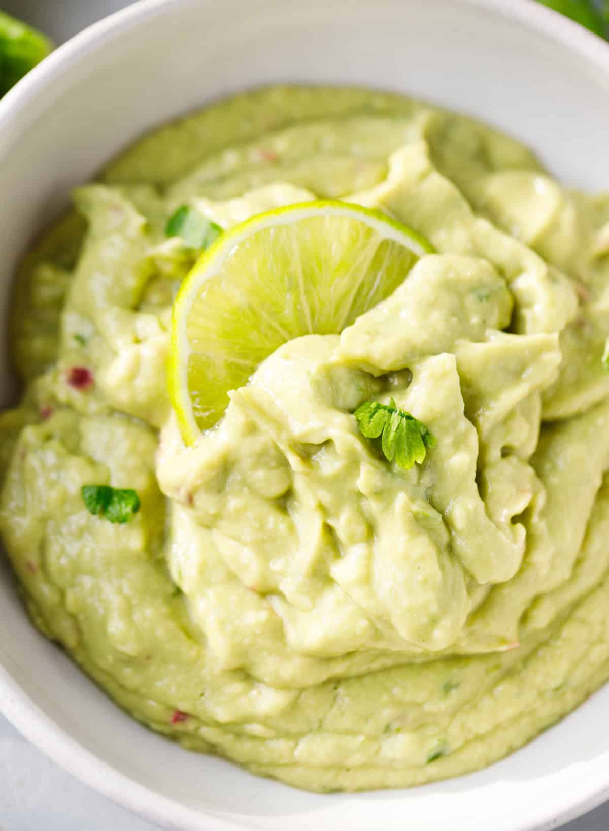 Close-up of smooth homemade blender guacamole topped with a lime slice and fresh cilantro.