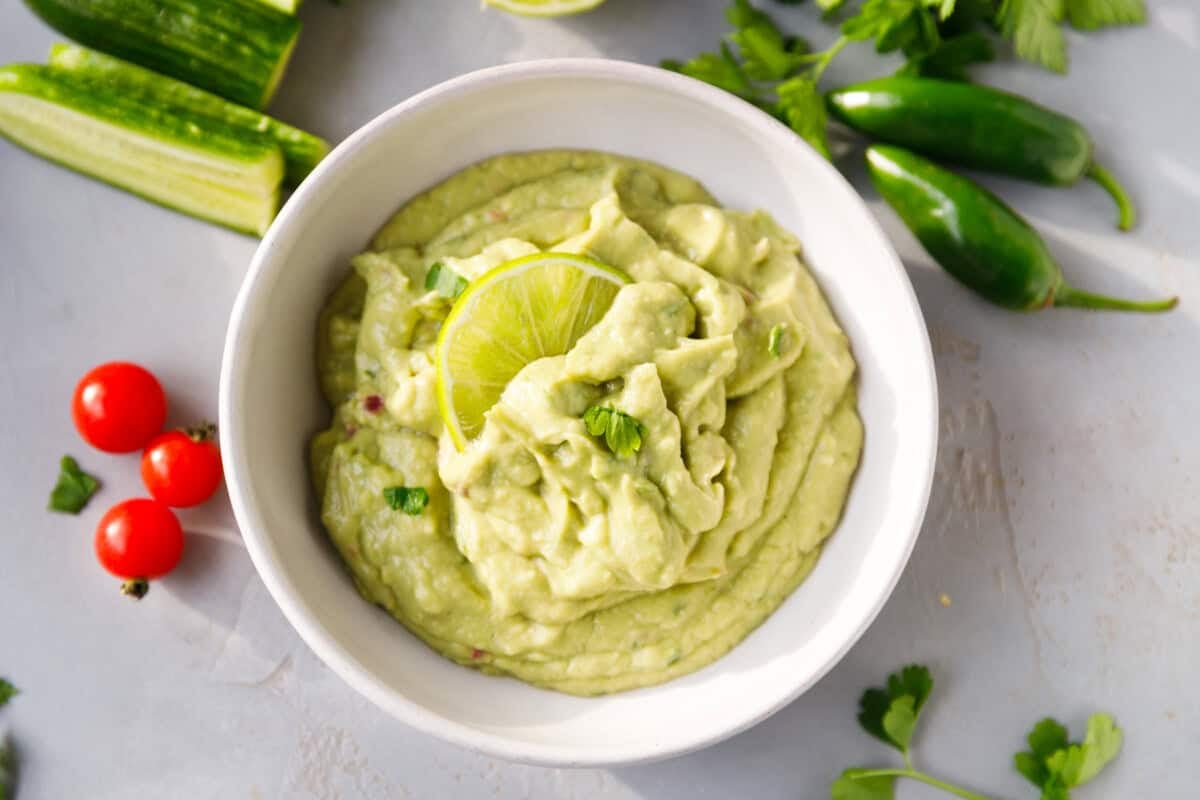 Bowl of creamy blender guacamole surrounded by cherry tomatoes, jalapeños and fresh cilantro.