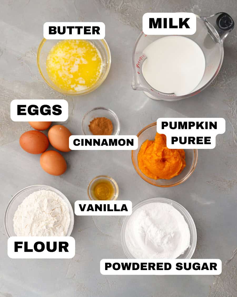 Ingredients for pumpkin magic custard cake including butter, milk, eggs, cinnamon, pumpkin puree, vanilla, flour, and powdered sugar arranged on a light gray surface.