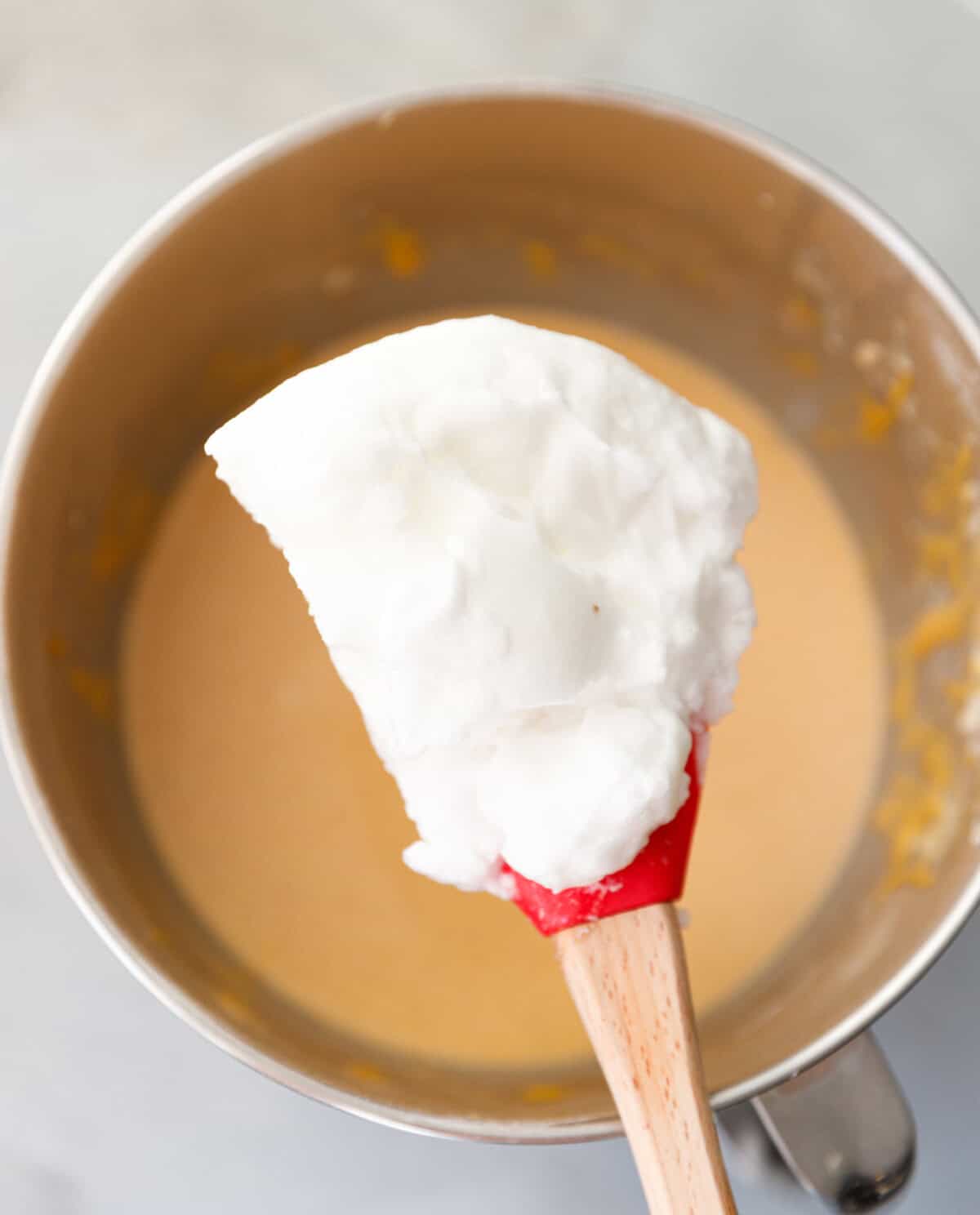 Spatula with whipped egg whites being added to pumpkin custard cake batter in a mixing bowl.