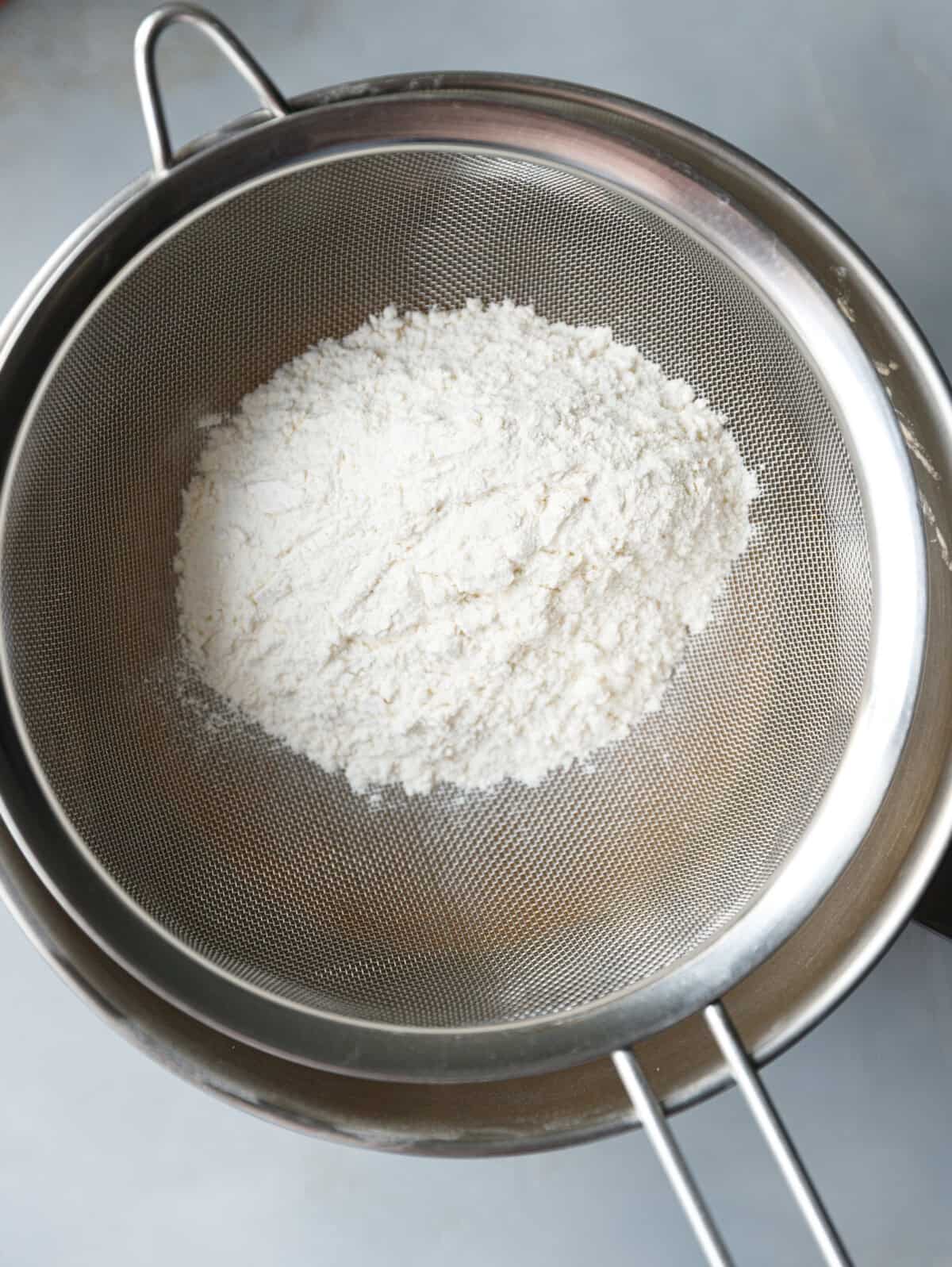 Sifting all-purpose flour through a fine mesh sieve into the pumpkin custard cake batter.
