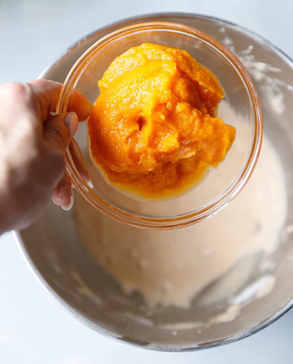 Hand holding a bowl of pumpkin puree over a mixing bowl to add into the custard cake batter.