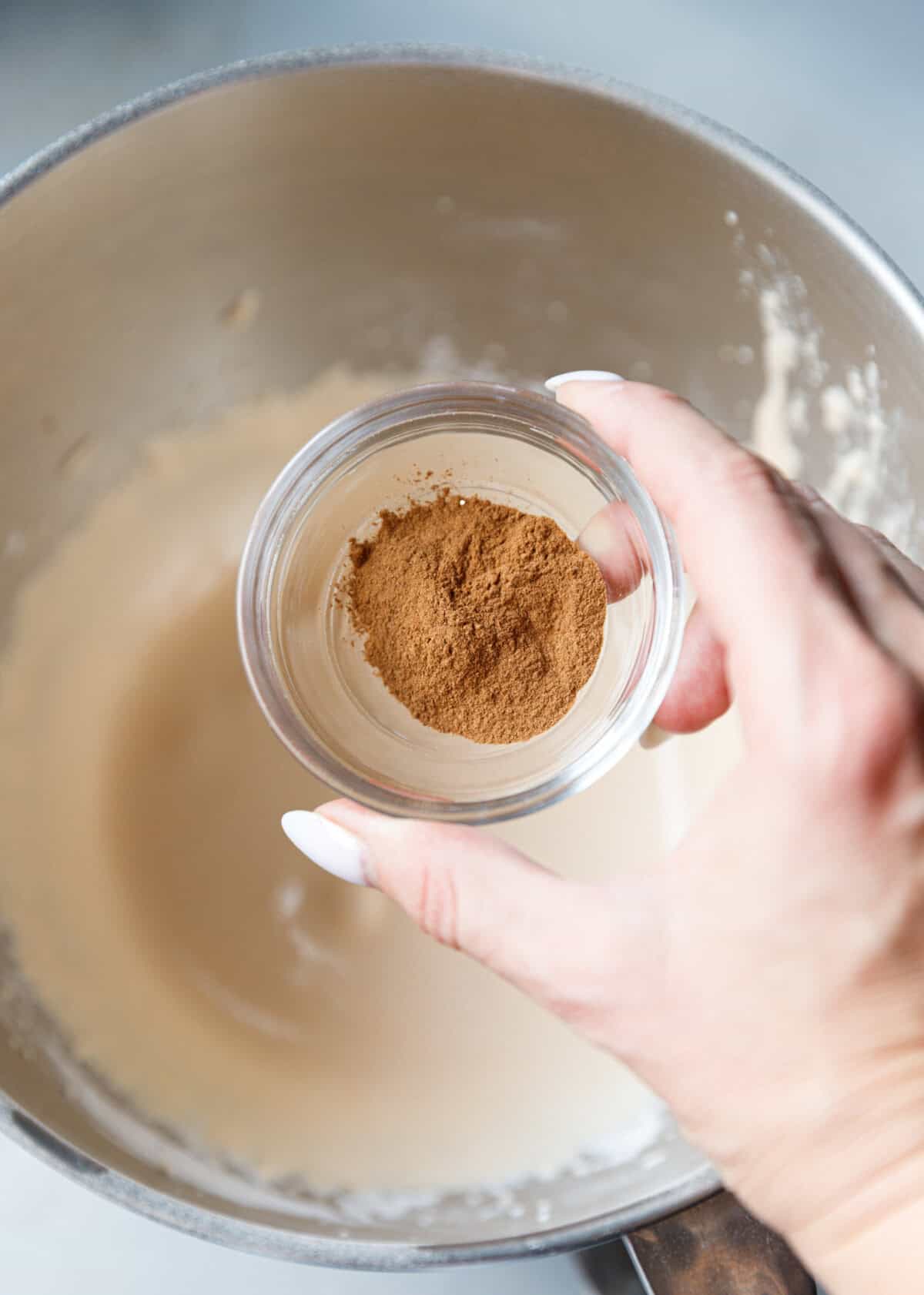Hand holding a small glass bowl of ground cinnamon above pumpkin custard cake batter.