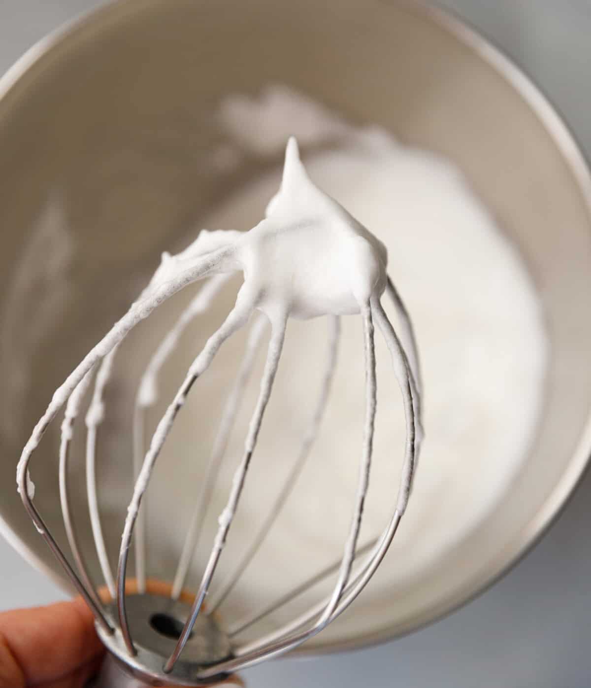 Hand holding a whisk attachment with stiff, glossy egg whites after whipping for pumpkin magic custard cake.