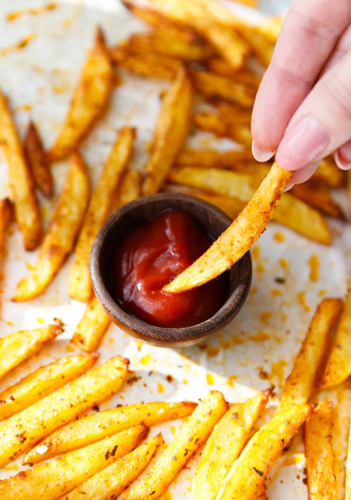 Hand dipping a crispy baked oven fry into a small bowl of ketchup.