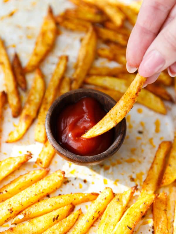 Hand dipping a crispy baked oven fry into a small bowl of ketchup.