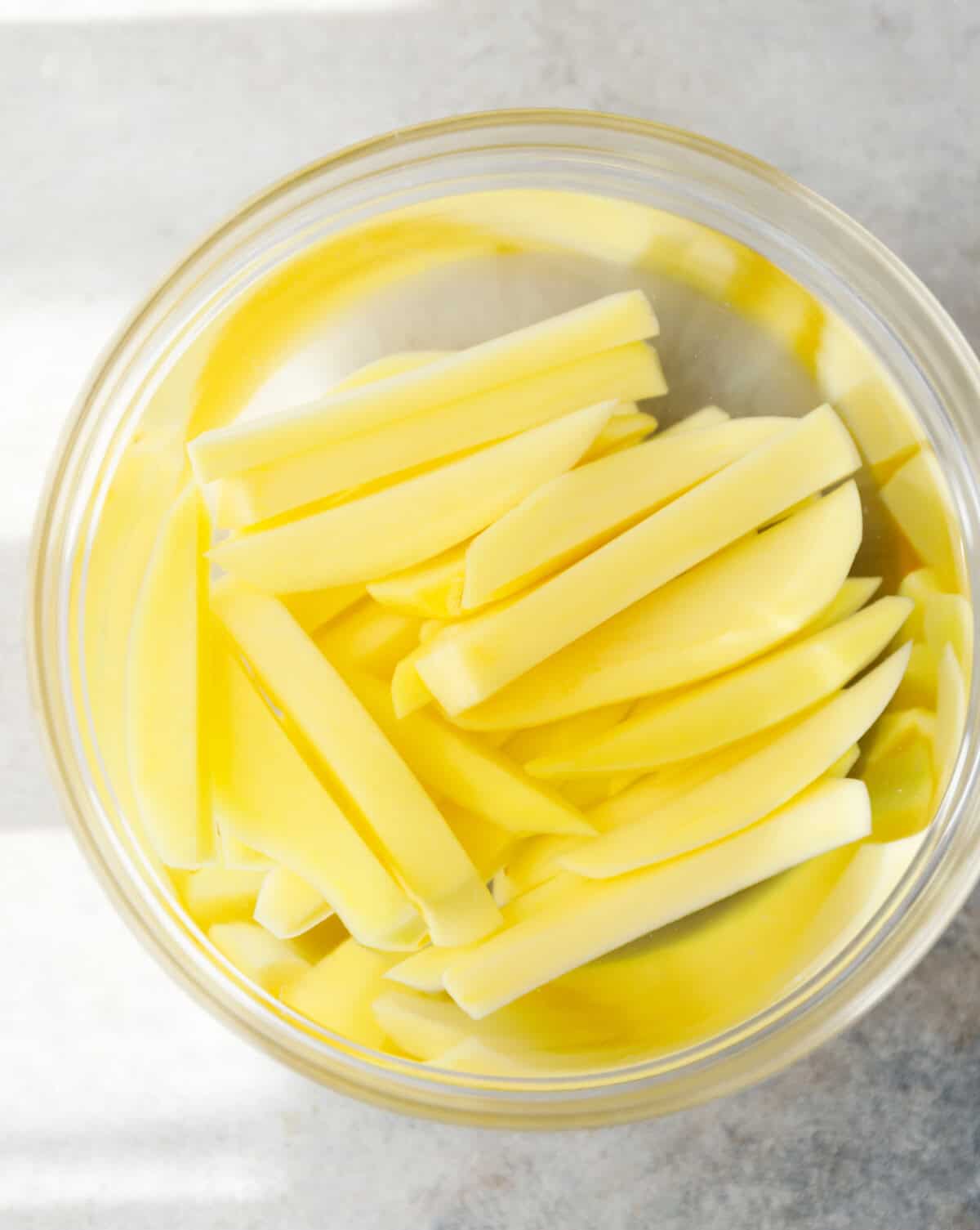 Potato matchsticks soaking in a glass bowl of cold water.