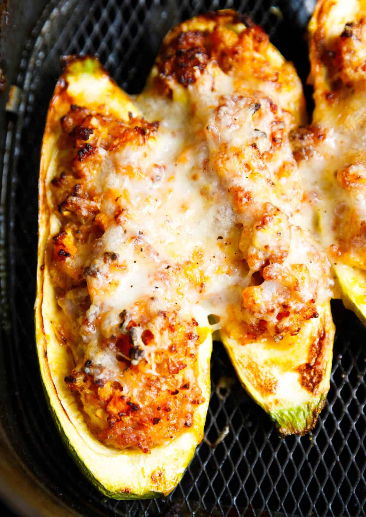 Stuffed zucchini boats in a black air fryer basket with melted cheese and lightly charred edges