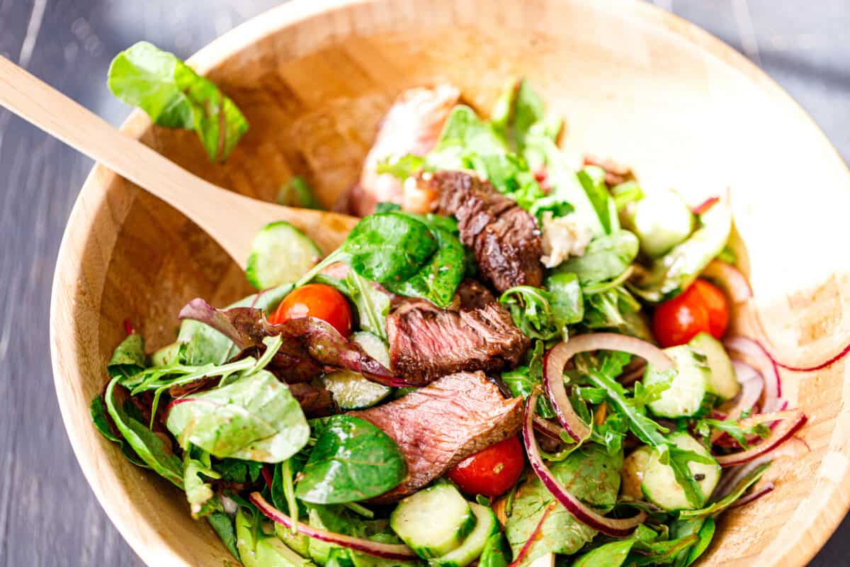 Freshly mixed steak salad served in a wooden bowl with colorful vegetables and sliced steak.