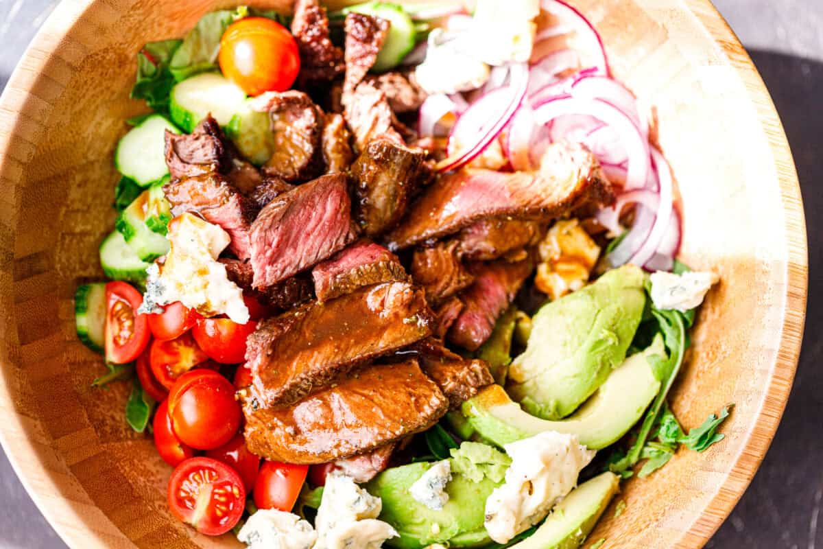 Warm sliced steak added on top of a fresh salad with tomatoes, cucumbers, avocado, and red onion.